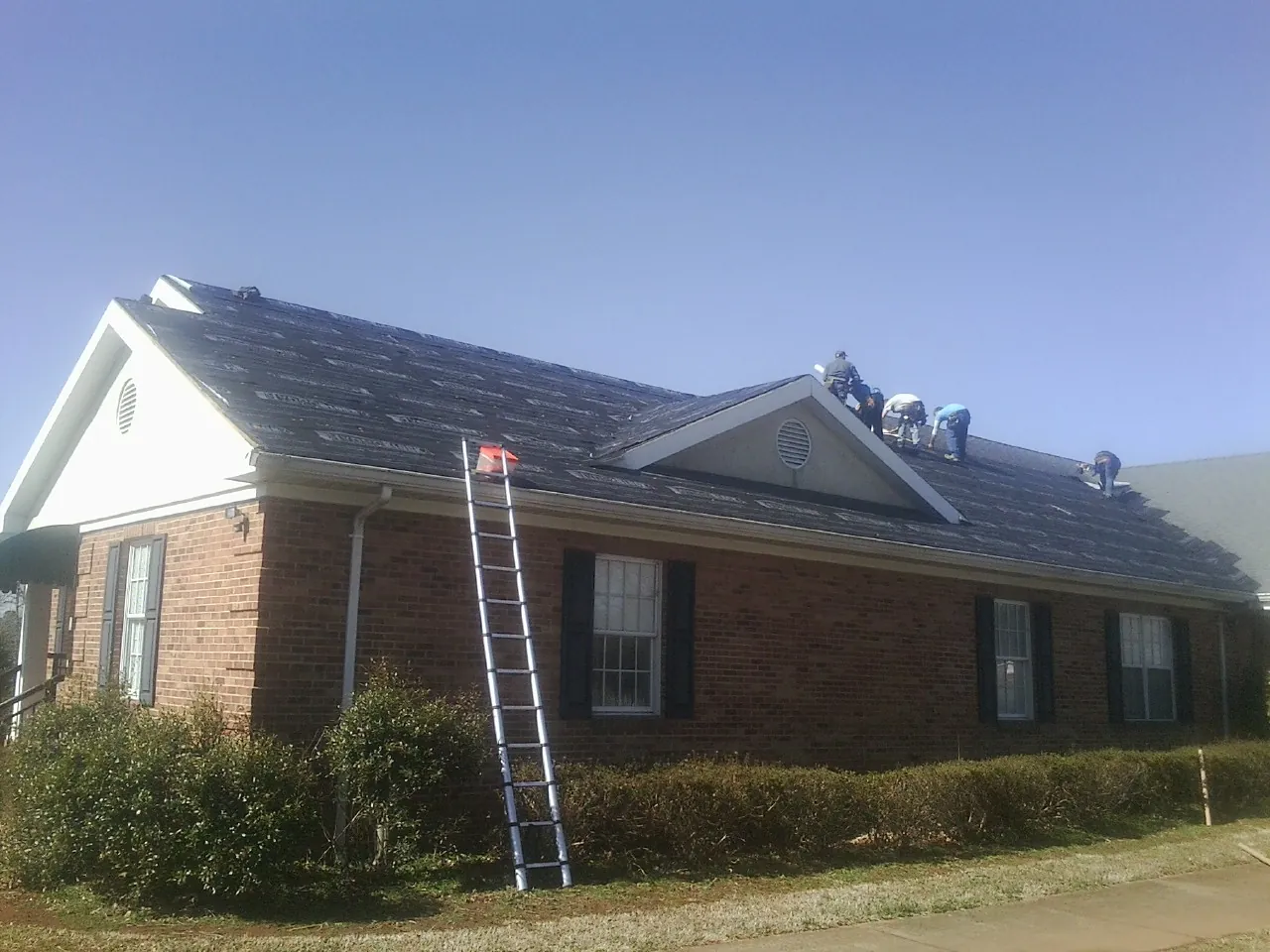 Crew tearing shingles off a brick ranch roof with a ladder set — Emergency Roof Repair work by our crew in Cashiers