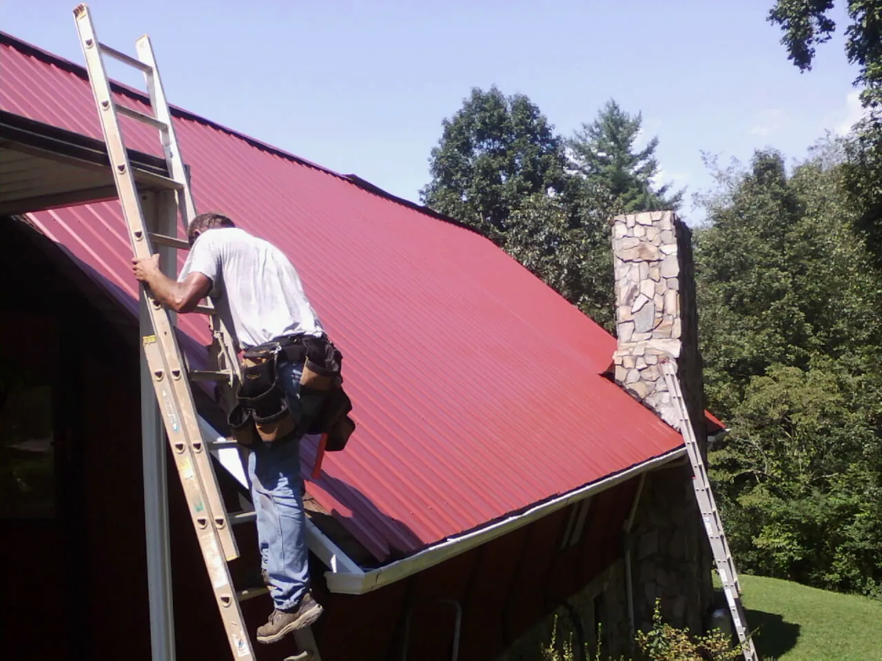 Roofer on a ladder beside a red metal roof and stone chimney — Soffit & Fascia Repair by Roofing Brothers in Cashiers